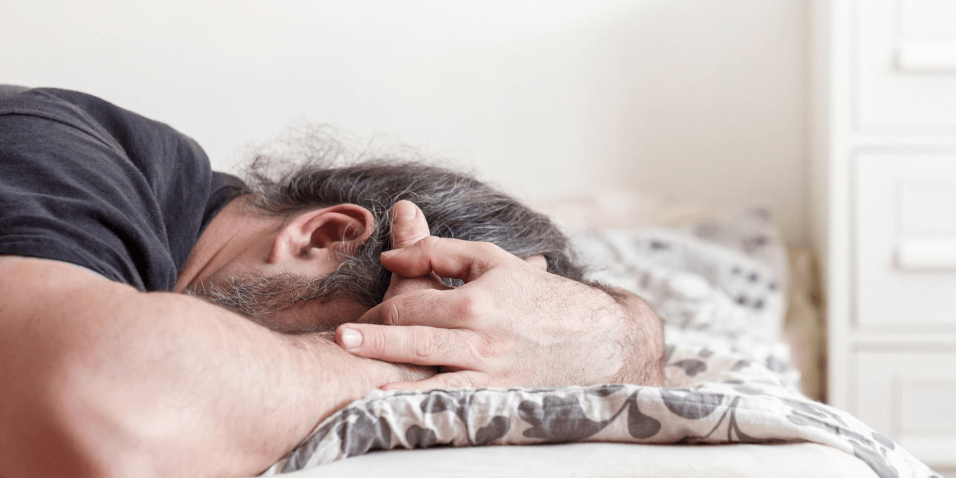 A man lying facedown on a bed in a brightly lit room. His arms are around his face.